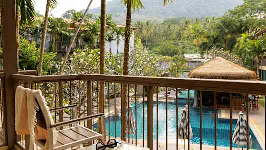 Utsikt fra en balkong over et basseng omgitt av palmer og grønn vegetasjon, med en stol og et bord i forgrunnen. (AI-generert tekst)