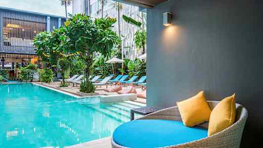 Moderne basseng med solsenger og vegetasjon. En sitteplass med puter i forgrunnen. (AI-generert tekst)