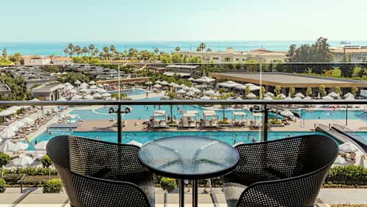 Utsikt fra en balkong med et bord og to stoler, med utsikt over et basseng og havet i bakgrunnen. (AI-generert tekst)