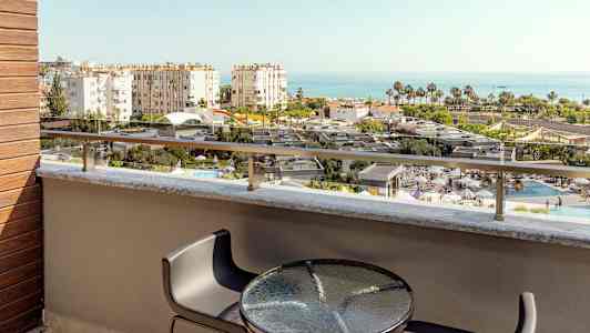 Utsikt fra en balkong med bord og stoler over et basseng og bygninger mot havet. (AI-generert tekst)