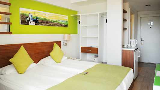 Hotellrom med en dobbeltseng, fargede puter, grønne dekorasjonsdetaljer og en åpen garderobe. (AI-generert tekst)