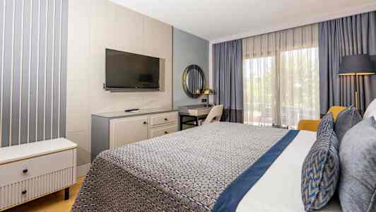 Hotellrom med moderne innredning, inkludert seng med blå detaljer, skrivebord, TV og store vinduer med gardiner. (AI-generert tekst)