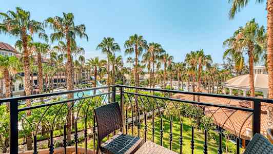 Utsikt fra en balkong over en hage med palmer og bygninger i bakgrunnen. (AI-generert tekst)