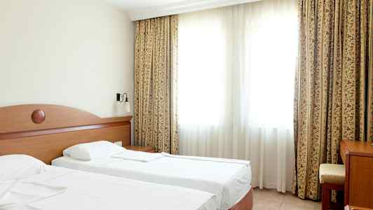 Et hotellrom med to enkeltsenger, et bord og en stol foran et stort vindu med gardiner. (AI-generert tekst)