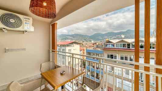 Balkong med et bord og to stoler, utsikt over bygninger og fjell. (AI-generert tekst)
