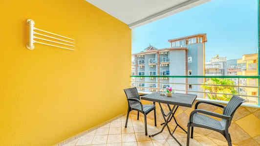 En balkong med en gul vegg, et bord med to stoler og utsikt over andre bygninger. (AI-generert tekst)