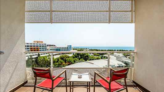 To røde stoler og et bord på en balkong med utsikt over havet og hotellbygninger. (AI-generert tekst)