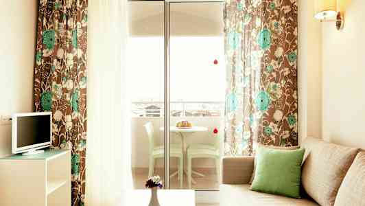 Sofa med grønn pute, TV og et bord med vase foran. Blomstrete gardiner og glassdør til en terrasse med bord og stoler. (AI-generert tekst)