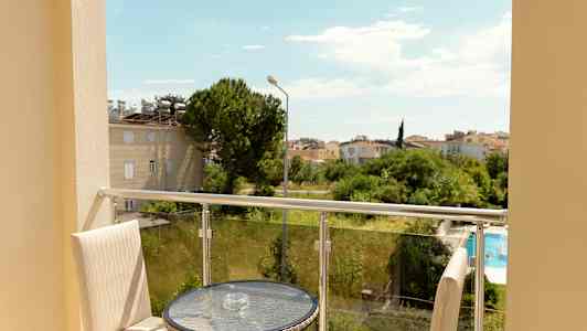 Utsikt fra en balkong med bord og stoler, mot grøntområder og et boligområde. (AI-generert tekst)