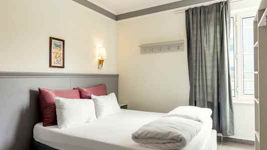 Bilde av et hotellrom med en dobbeltseng, dekorert med sengetøy i hvitt og rødt samt en lys grå sengegavl. (AI-generert tekst)