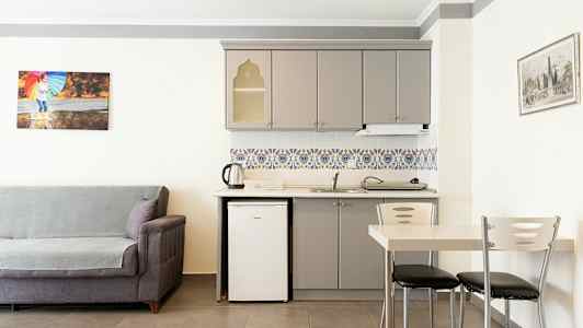 Et rom med grå sofa, en kjøkkenkrok med hvit kjøleskap, grå skap og fargerike fliser. En spisestue med bord og stoler står ved siden av. (AI-generert tekst)