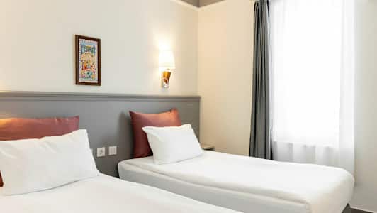 To senger med hvitt sengetøy og røde puter står i et rom med lyse vegger og en vegglampe. Et vindu med hvite gardiner er til høyre. (AI-generert tekst)