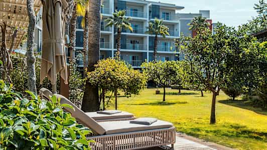 To solsenger på en terrasse med utsikt over en plen og trær, med et leilighetshotell i bakgrunnen. (AI-generert tekst)