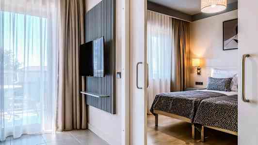 Moderne hotellrom med to enkeltsenger, en flatskjerm-TV på veggen og store vinduer med gardiner. (AI-generert tekst)