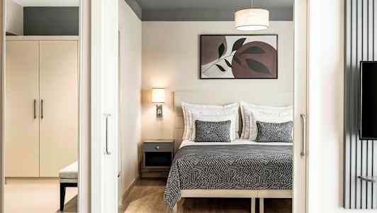 Et soverom med en dobbeltseng med grått sengeteppe og matchende puter, en vegglampe og et maleri over sengen. Et skap og benk vises i forgrunnen. (AI-generert tekst)