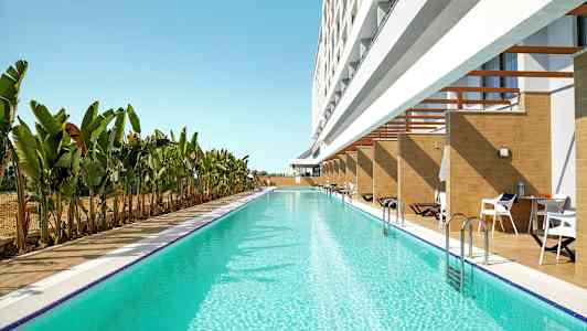 Lang basseng med klart blått vann ved siden av en bygning med private terrasser og solsenger, omgitt av banantrær. (AI-generert tekst)