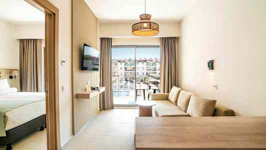 Et hotellrom med sittegruppe, seng og TV, samt balkong med utsikt over bassengområdet. (AI-generert tekst)