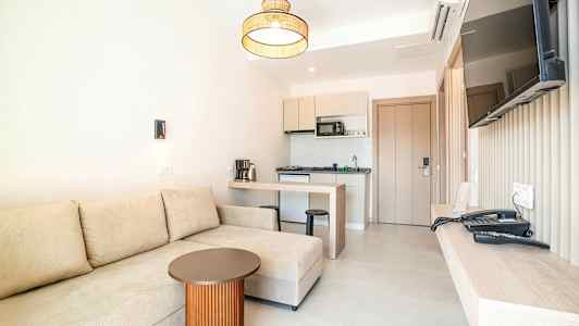 Et hotellrom med en beige sofa, et lite bord, et kjøkken og en veggmontert TV. (AI-generert tekst)