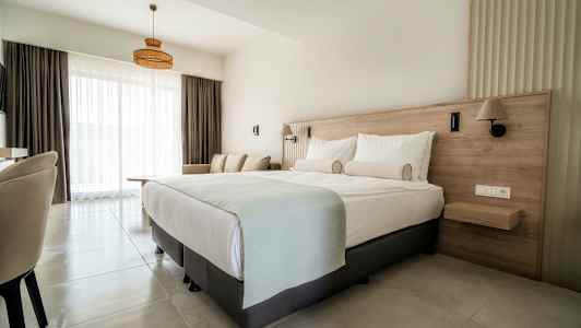 Et hotellrom med en dobbeltseng, sofa og store vinduer med lyse gardiner. (AI-generert tekst)