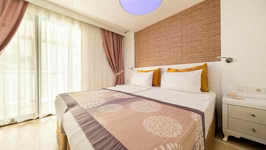 Hotellrom med en dobbeltseng, dekorert med mønstret sengeteppe og puter. Vindu med gjennomsiktige gardiner og lilla draperier i bakgrunnen. (AI-generert tekst)