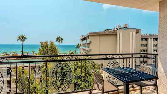 Utsikt over en balkong med bord og stoler, samt hav og palmer i bakgrunnen. (AI-generert tekst)