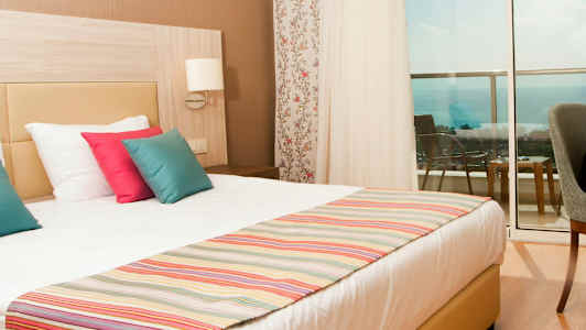 Hotellrom med en dobbeltseng, fargerike puter og utsikt mot havet fra balkongen. (AI-generert tekst)