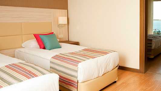 To enkeltsenger med stripete sengetepper og fargerike puter, i et rom med tregulv og lyse vegger. (AI-generert tekst)