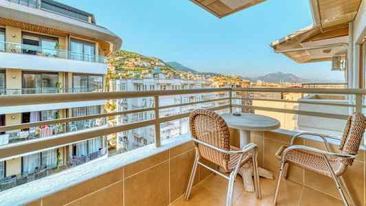 Balkong med to stoler og et bord, med utsikt over bygninger og fjell. (AI-generert tekst)