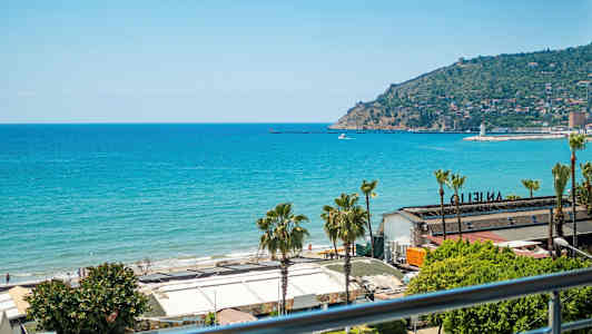 Utsikt over havet og en strand med bygninger i forgrunnen. En odde med bebyggelse er synlig i bakgrunnen. (AI-generert tekst)