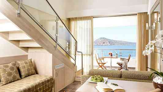 En lys stue med utsikt over havet, en balkong med sittemøbler og et salongbord med druer og en bok. (AI-generert tekst)