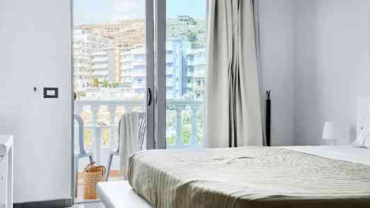 Utsikt fra et hotellrom, med seng og en åpen balkongdør. Balkong med stoler og utsikt over bygninger. (AI-generert tekst)