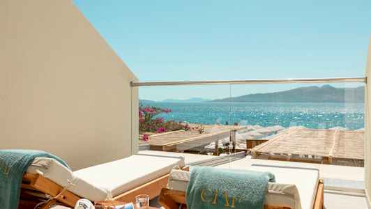 En solseng med et grønt håndkle ved en balkong med glassrekkverk, med utsikt over havet og en strand med parasoller. (AI-generert tekst)