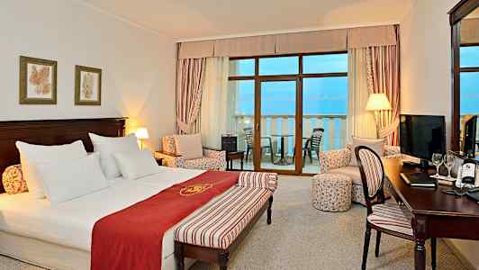 Et hotellrom med dobbeltseng, sittegruppe og balkong med havutsikt. (AI-generert tekst)