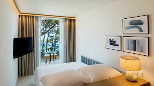 Et hotellrom med en seng, en lampe på bordet, veggdekorasjoner og et vindu som vender mot havet og trær. (AI-generert tekst)