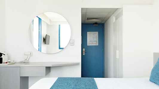 Et hotellrom med en blå dør, et rundt speil, en TV og en seng med blå detaljer. (AI-generert tekst)