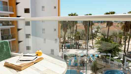 En balkong med en åpen bok, et par briller og et eple på et bord. I bakgrunnen vises et basseng med omkringliggende palmer og bygninger. (AI-generert tekst)