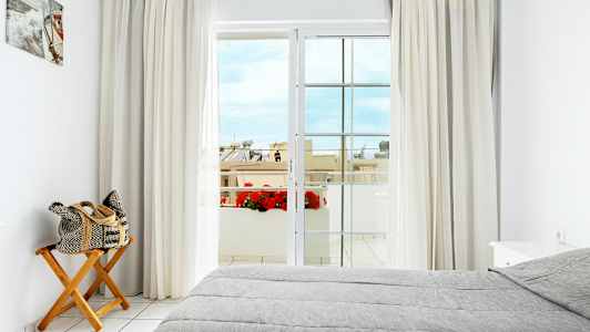 Et lyst soverom med en grå oppredd seng, en trestol med en sammenrullet veske, gardiner og en balkongdør som leder til en utsikt med blomster og bygninger. (AI-generert tekst)