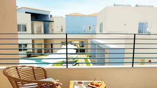 Utsikt fra en balkong mot en gårdsplass med et lite basseng, omgitt av lyse bygninger. På balkongen er det en stol og et bord med solbriller og en kaffekopp. (AI-generert tekst)