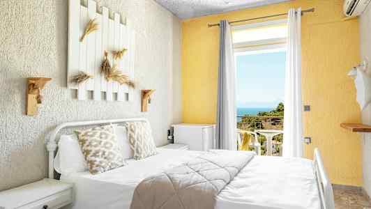 Lyst hotellrom med seng, dekorerte puter og utsikt mot balkong med hav og grøntområder. (AI-generert tekst)