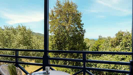 Utsikt fra en balkong med bord og stoler mot trær og hav i bakgrunnen. (AI-generert tekst)