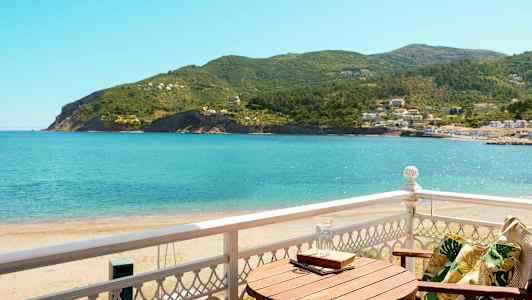 Utsikt fra en balkong mot havet og frodige åser, med en trestol og et bord. (AI-generert tekst)