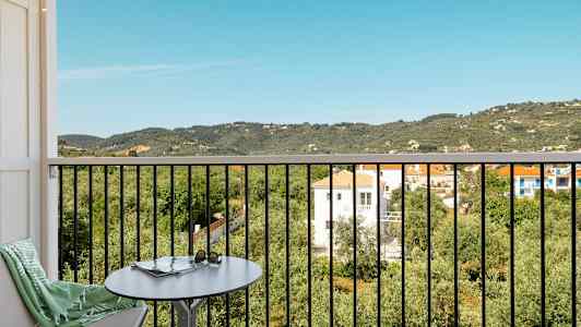 Utsikt fra en balkong med bord og stol over et frodig landskap med hus og fjell. (AI-generert tekst)