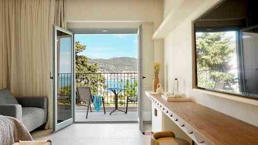 Et hotellrom med åpen balkongdør som viser utsikt over havet og omkringliggende grøntområder. (AI-generert tekst)