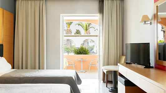 Hotellrom med to senger, skrivebord, TV og en sittegruppe ved et vindu som vender mot en terrasse med utsikt over palmer. (AI-generert tekst)