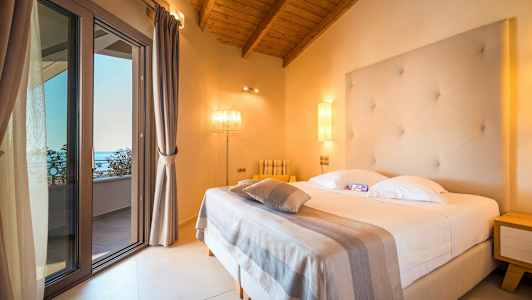 Et hotellrom med en seng, en strukturert hvit sengegavl og en taklampe. Glassdøren fører til en balkong. (AI-generert tekst)