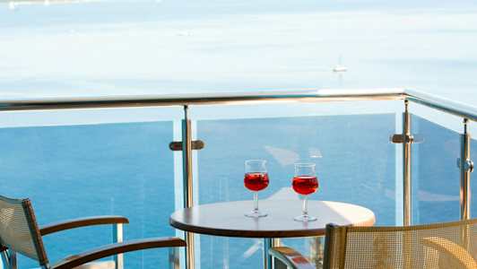 To glass med rød drikk på et bord på en balkong med utsikt over havet. (AI-generert tekst)