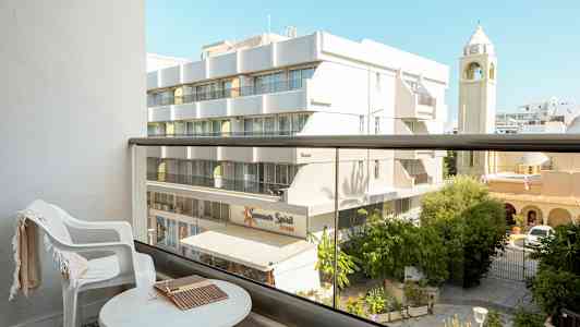 Utsikt fra en balkong med et bord og en stol, med en bygning og et kirketårn i bakgrunnen. (AI-generert tekst)