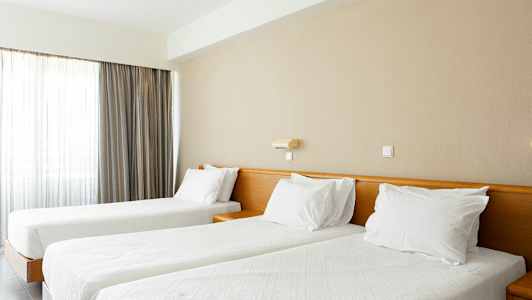 To senger med hvite duker og puter i et lyst rom med gardiner. (AI-generert tekst)