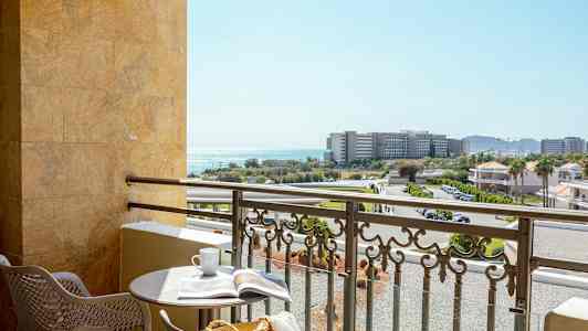 En balkong med et rundt bord, to stoler og utsikt over bygninger, veier og havet. (AI-generert tekst)