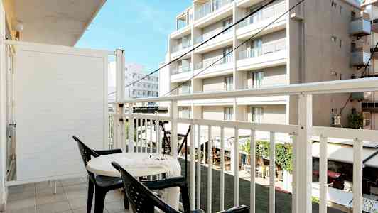 En balkong med to stoler og et bord, utsikt mot bygninger. (AI-generert tekst)